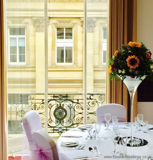 Tall martini glass table centrepieces on mirror plates with sunflowers, yellow gerberas, white spray roses, purple lissianthus at Doubletree by Hilton Liverpool